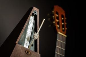 Color shot of a vintage metronome, next to an acoustic guitar, on a black background.